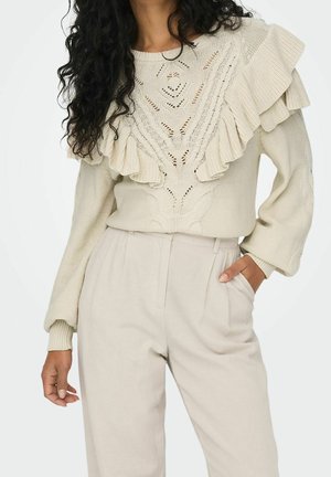 Knit sweater in light beige with a textured pattern and prominent ruffle detailing on the shoulders. Paired with loose-fitting beige trousers.