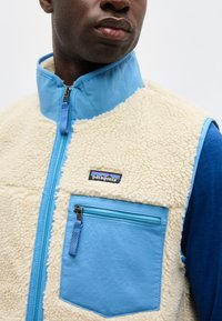 Vīrietis valkā krēmkrāsas sherpa vilnas vestu ar zilu apkakli, rāvējslēdzēja kabatu un Patagonia logotipu uz krūtīm, pāri zilai garroku krekla.