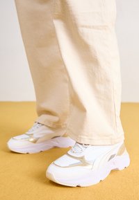 Weiße Sneakers mit metallischen Goldakzenten, ausgestattet mit einer klobigen Sohle und Mesh-Stoff. Getragen mit leicht beigen, locker sitzenden Hosen auf einer gelben Fläche.