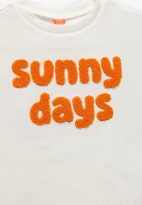 Hvid tekstureret stof med fluffy orange bogstaver, der står "solrige dage" i en legesyg skrifttype. Mærke synligt ved halsåbningen.