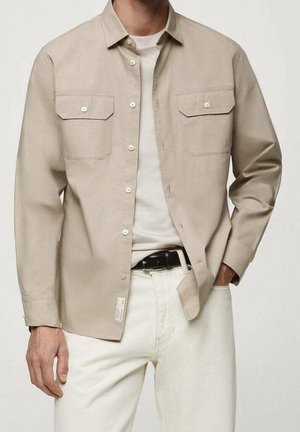 Muž v světlé béžové košili na knoflíky přes bílé tričko, zastrčené do krémových kalhot s černým opaskem, rukou v kapse.