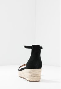 Black suede wedge sandal with a braided jute platform, ankle strap with metal buckle, and open toe design.