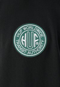 Črna tkanina vsebuje okroglo zeleno in belo vezeno nalepko z besedilom "HUF WORLDWIDE" in "TRANSIT AUTHORITY".