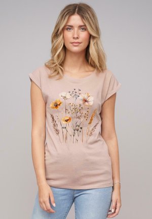 T-shirt à manches courtes rose clair avec un imprimé floral dans des teintes pastel et dorées, mettant en avant diverses fleurs sauvages et herbes.