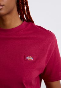 Lähedalne foto inimesest, kes kannab burgundi värvi Dickies T-särki, punase punutud juustega vasakul pool, lihtsa tausta ees.