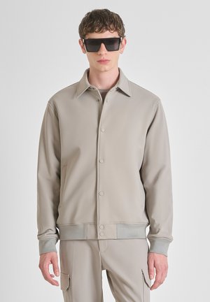 Giacca beige chiaro in un materiale liscio e elasticizzato. Presenta un colletto a punta, bottoni a pressione, tasche laterali e polsini a costine.