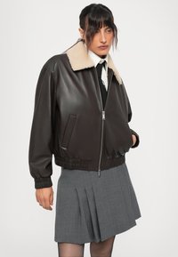 Person wearing dark brown leather jacket with beige shearling collar, white shirt, black tie, and pleated gray skirt, standing against white background.