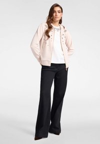 Chaqueta con cremallera de color rosa claro con adornos florales, combinada con una blusa blanca y pantalones de pierna ancha negros. Tejido suave, diseño casual.