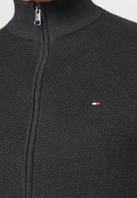 Primo piano di un maglione zip-up scuro e Texturato con un piccolo logo rettangolare rosso, bianco e blu sul petto vicino alla cerniera.