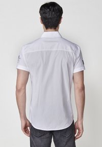 Camisa blanca de manga corta con un cuello casual. Presenta una textura suave y detalles de costura. Combinada con jeans oscuros, mostrando un diseño limpio.