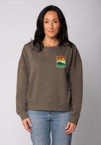 Vrouw met donker haar draagt een bruine sweatshirt met een "Good Vibe" patch van een berg en zon, en lichte blauwe spijkerbroek, staande voor een effen achtergrond.