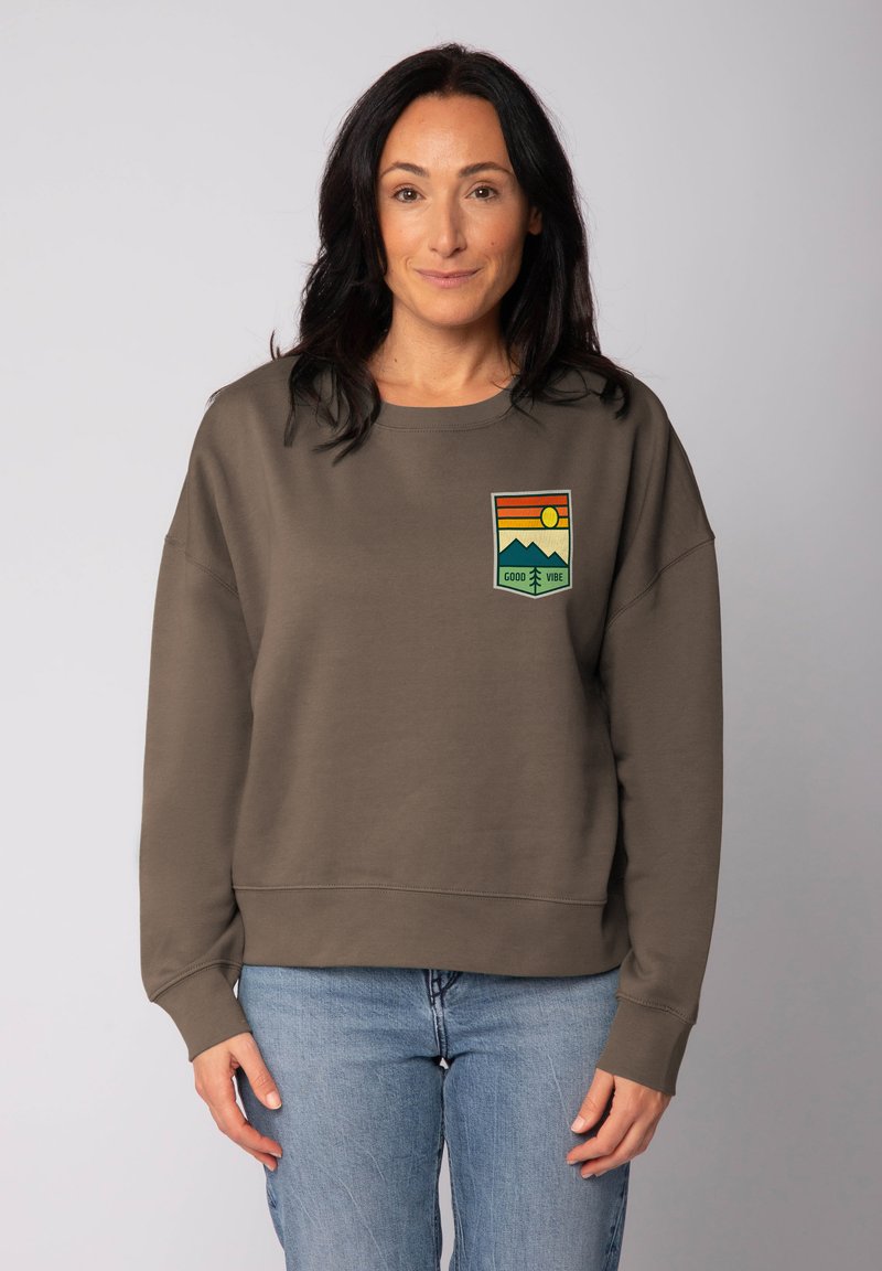 Vrouw met donker haar draagt een bruine sweatshirt met een "Good Vibe" patch van een berg en zon, en lichte blauwe spijkerbroek, staande voor een effen achtergrond.