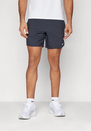 Mężczyzna w czarnych sportowych szortach Nike, białych skarpetkach z logo Nike oraz białych butach do biegania, stojący na jednolitym tle.