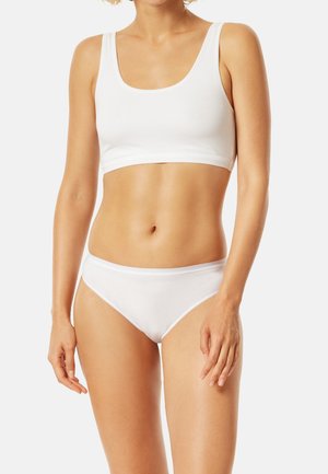 Woman wearing plain white sports bra and matching white underwear, standing against a neutral background.