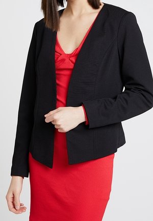 Vrouw draagt een nauwsluitend zwart colbert over een rode geribbelde jurk met een gedraaid detail bij de halslijn, staand tegen een effen achtergrond.
