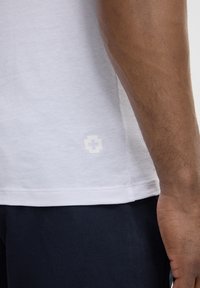 Witte t-shirt met korte mouwen, gemaakt van lichte stof, met een subtiele logo-detail aan de onderzijde. Draagt met donkere broek.