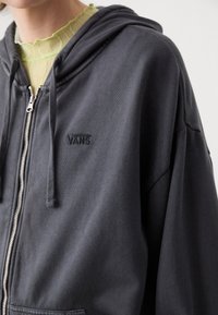 Personne portant un sweat à capuche Vans zippé gris foncé par-dessus un haut vert clair à volants et col montant, coupé au niveau du cou et des épaules.