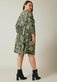 Vestido verde con estampado de paisley, de mangas largas y cinturilla anudada, combinado con botas negras hasta el tobillo con un tacón grueso y herrajes dorados.