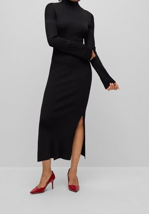 Nainen pukeutuneena pitkään mustaan ribbineulemekkoon, jossa on sivuhalkio, ja punaisiin kärjestä teräviin korkeisiin korkokenkiin, yksinkertaisen harmaan taustan edessä.