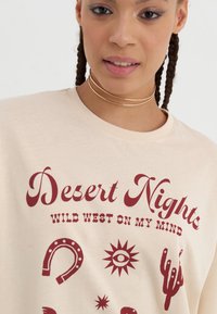 Beige t-shirt met korte mouwen met maroon graphics, waaronder de tekst "Desert Nights" en verschillende westerse iconen zoals cactussen en hoefijzers.
