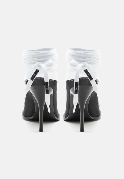 Chaussures à talons hauts noires avec des liens blancs noués autour de la cheville portant le texte "MOSCHINO", vues de dos sur un fond blanc.