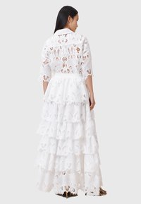 Biała sukienka maxi z koronki, z wielowarstwowymi falbanami, kołnierzykiem i rękawami do łokcia, ozdobiona kwiatowymi wzorami i przezroczystą fakturą.