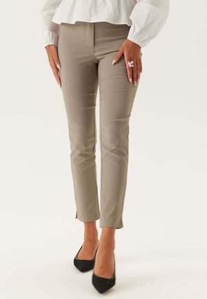 Pantalons slim beige avec une texture lisse, présentant une taille basse et un ourlet fendu court. Associés à des talons noirs pointus.