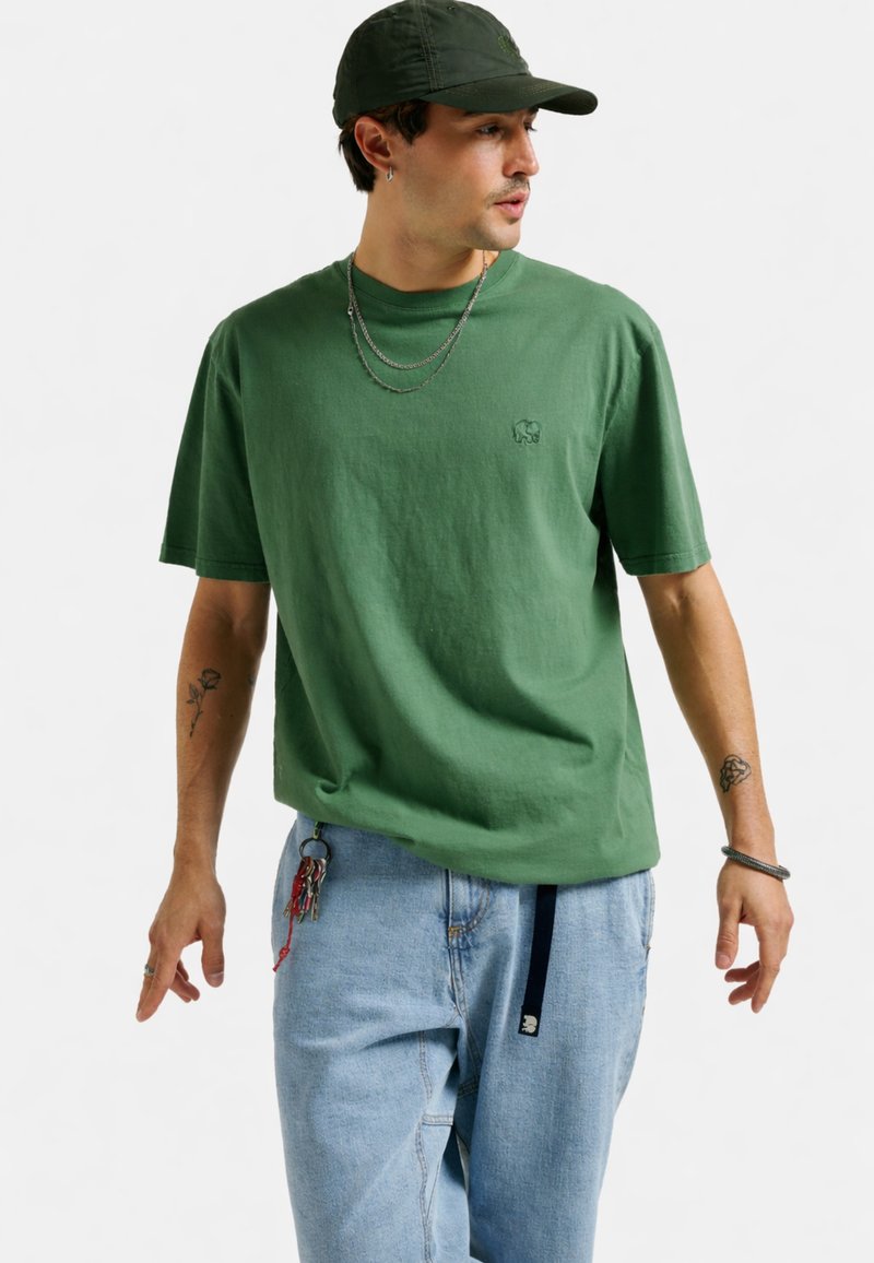 Uomo che indossa un berretto verde, una maglietta verde con un piccolo logo a forma di elefante, catene a strati, jeans azzurri con chiavi appese e braccia tatuate.