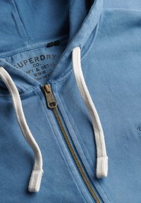 Felpa con zip di colore azzurro chiaro realizzata in tessuto morbido. Presenta corde bianche, una zip dorata e un logo sul davanti.