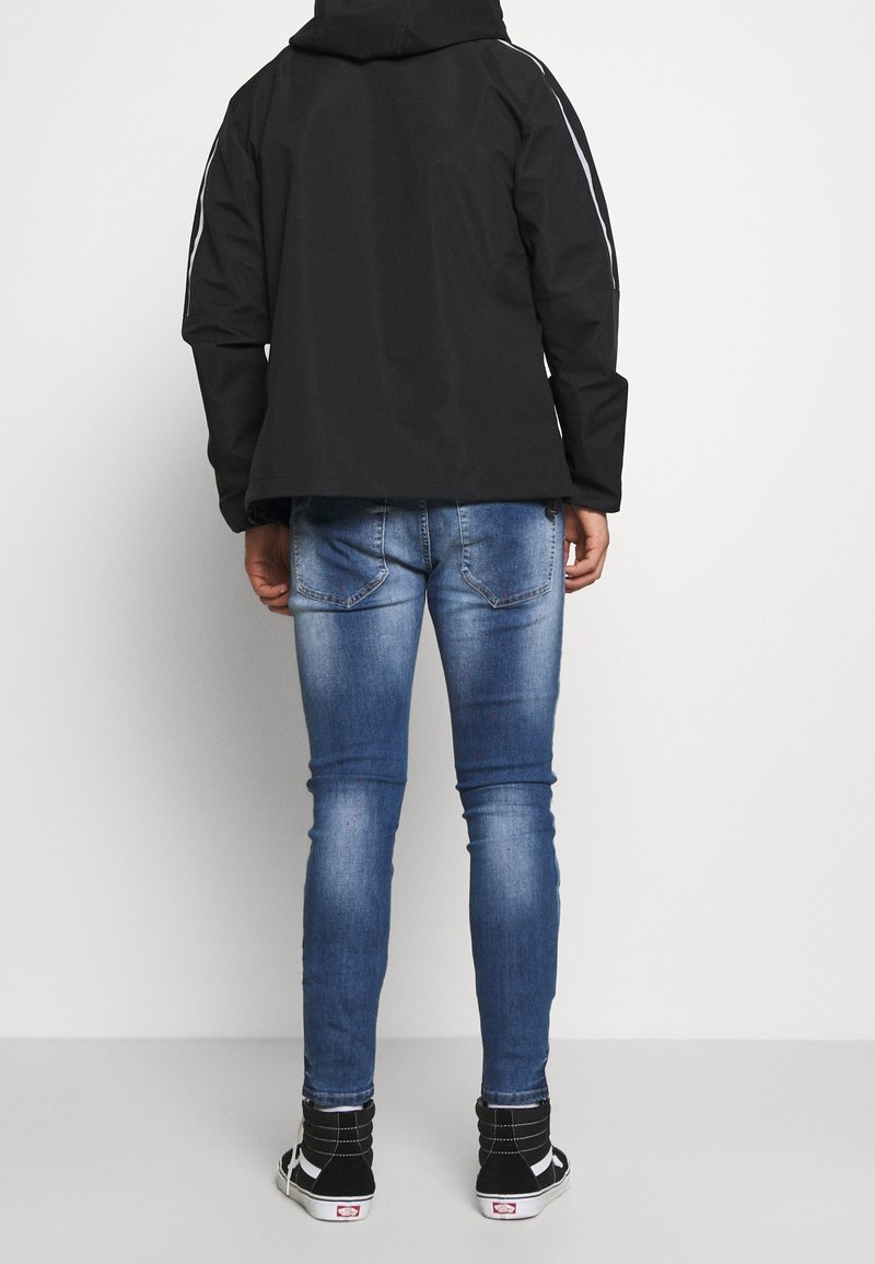 Chaqueta negra con capucha y rayas laterales, combinada con jeans azules. Los jeans presentan un patrón desgastado y son ajustados. Zapatillas negras con acentos blancos.