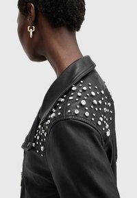 Black leather jacket with silver studded embellishments on the shoulder. Features a classic collar and zip closure. Smooth texture.