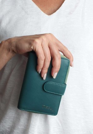 Portafoglio in pelle teal con una superficie liscia, dotato di chiusura a bottone e logo embossato. Tenuto in mano su uno sfondo in tessuto chiaro.