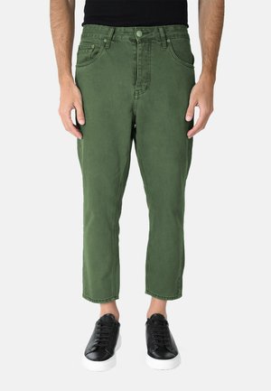 Uomo che indossa pantaloni corti verde oliva e scarpe da ginnastica nere con suole bianche, in piedi davanti a uno sfondo bianco uniforme.
