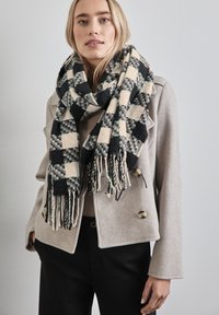 Vävd scarf i svart, kräm och grön rutmönster, med fransiga kanter. Modellen bär en ljusgrå jacka med knappdetaljer.