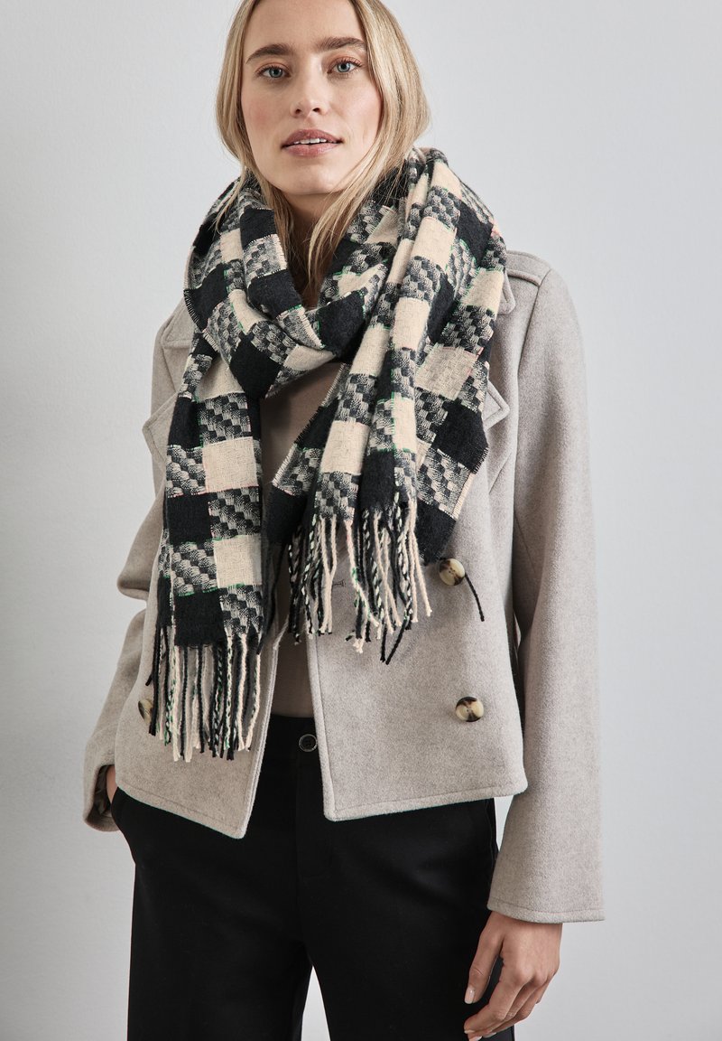Gewebter Schal in schwarz, creme und grünem Karomuster, mit fransigen Kanten. Das Model trägt eine hellgraue Jacke mit Knopfdetails.
