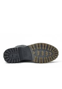 Suela de bota de goma negra con surcos profundos, que cuenta con una superficie texturizada y un logotipo rectangular. Patrón aserrado distintivo para tracción.
