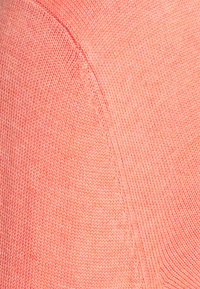 Sweter dzianinowy w koralowym kolorze, o gładkiej fakturze i zaokrąglonych szwach na ramionach. Widoczne przeszycia dodają tkaninie subtelnych detali.