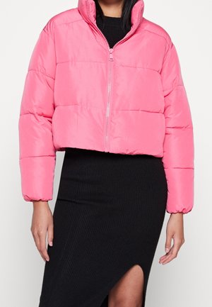 Vrouw draagt een cropped felroze donsjack met rits en een zwarte geribbelde rok met frontsplit tegen een witte achtergrond.