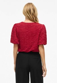 Haut court à manches courtes texturé rouge avec un motif de vagues en relief. Porté avec un pantalon noir, affichant une coupe décontractée et un col rond.