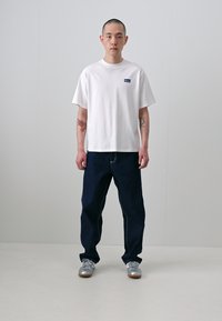 T-shirt branca de algodão com mangas curtas e um patch de logótipo azul, combinada com jeans azul escuro e ténis prateados.
