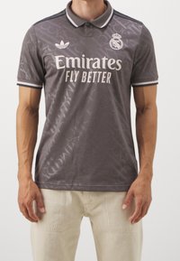 Mężczyzna ubrany w szary trykot piłkarski Real Madryt z logo Emirates Fly Better i beżowe spodnie, stojący na tle białym.