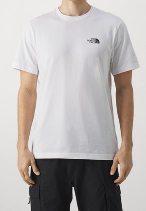 Mand iført en ensfarvet hvid kortærmet t-shirt med lille sort The North Face-logo på brystet og sorte cargobukser, stående mod hvid baggrund.