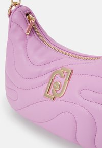 Sac à main en cuir lilas avec fermeture éclair dorée et détail décoratif de logo doré sur le côté, avec des motifs cousus ondulés.