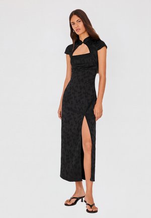 Femme portant une robe midi noire à motifs avec des manches gigot, un décolleté carré, une fente latérale haute et des sandales noires à talons.
