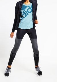 Zwarte leggings met een gestructureerd patroon en drie strepen op het onderbeen, gecombineerd met een lichtblauw T-shirt met een groot logo en een zwarte jas met ritssluiting.