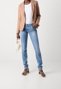 Jean bleu clair à coupe slim avec une texture délavée, associé à un blazer beige porté sur un haut blanc, tenant un petit sac à main crème.