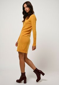 Abito in maglia giallo con maniche lunghe e design aderente. Indossato con stivali alla caviglia bordeaux con finitura lucida e tacco a blocco.