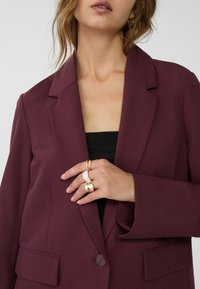 Bordeauxblazer met een gestructureerd ontwerp, metgeligde revers, enkele knoopsluiting en zijzakken; gedragen over een zwart top met ringen aan de hand.