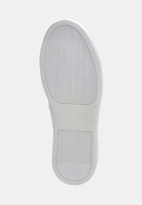 Semelle de chaussure de sport en caoutchouc blanc avec un motif ondulé texturé et une indentation rectangulaire. Logo "GUESS" embossé sur la zone du talon.
