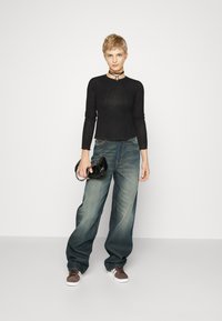 Schwarzes geripptes Langarmtop, weite blaue Jeans und braune Sneakers. Hält eine glänzende schwarze Handtasche mit silberner Kettendetail.